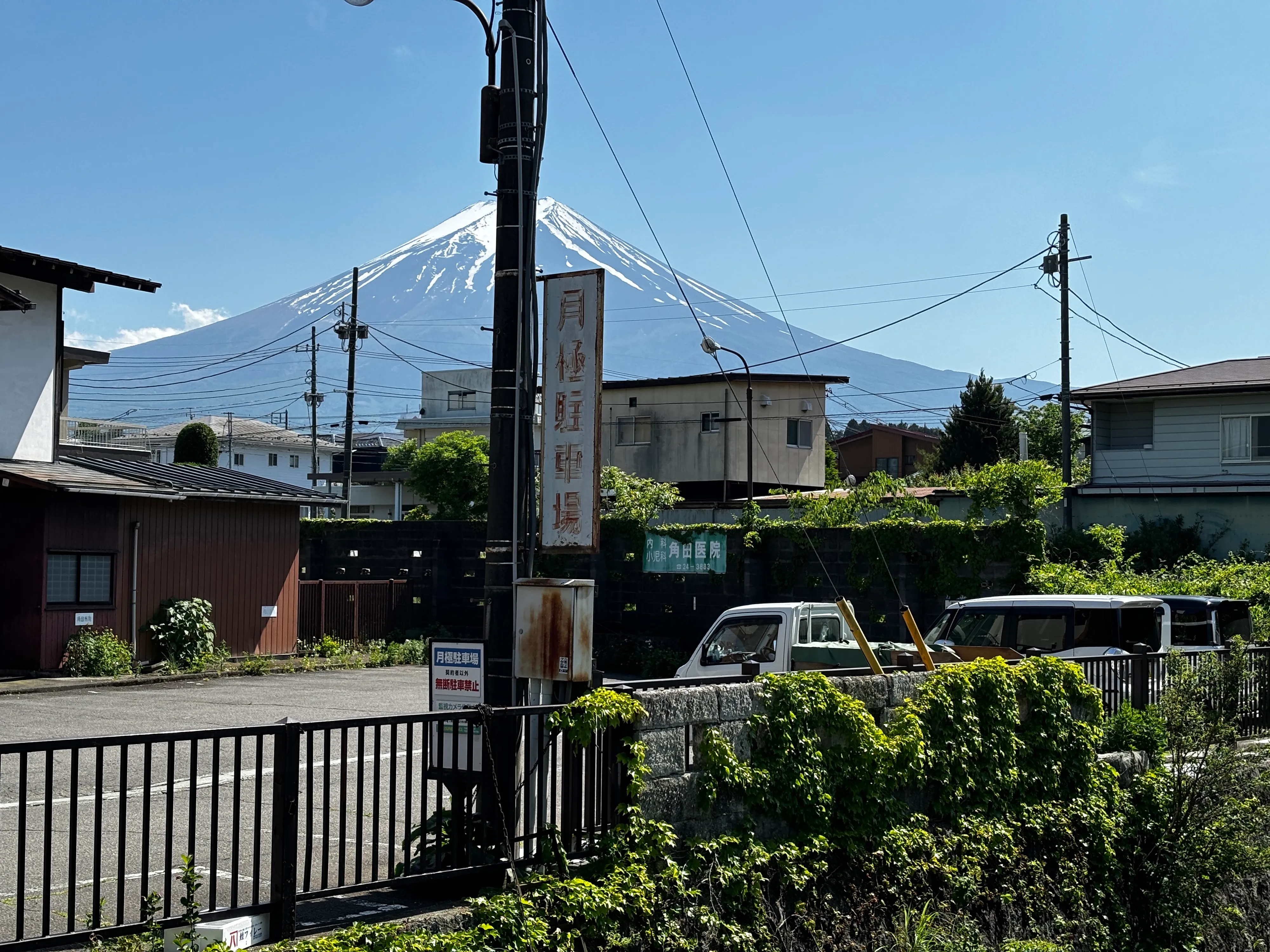 Snow-capped Mount Fuji rising over a residential street with power lines, a vertical shop sign and parked vans under a clear blue sky