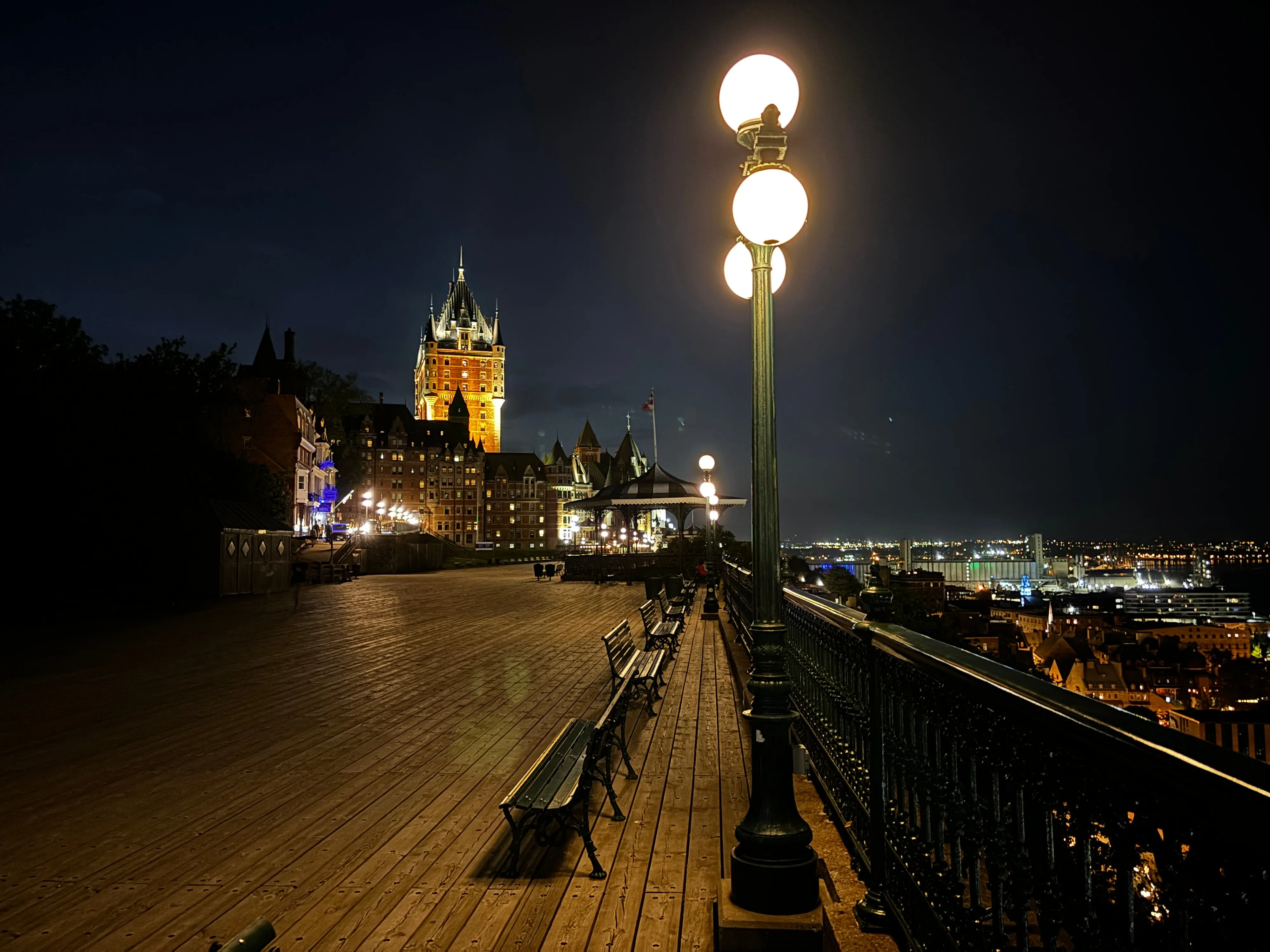 The lit towers of the Château Frontenac beyond an empty boardwalk, benches and a lamppost at night