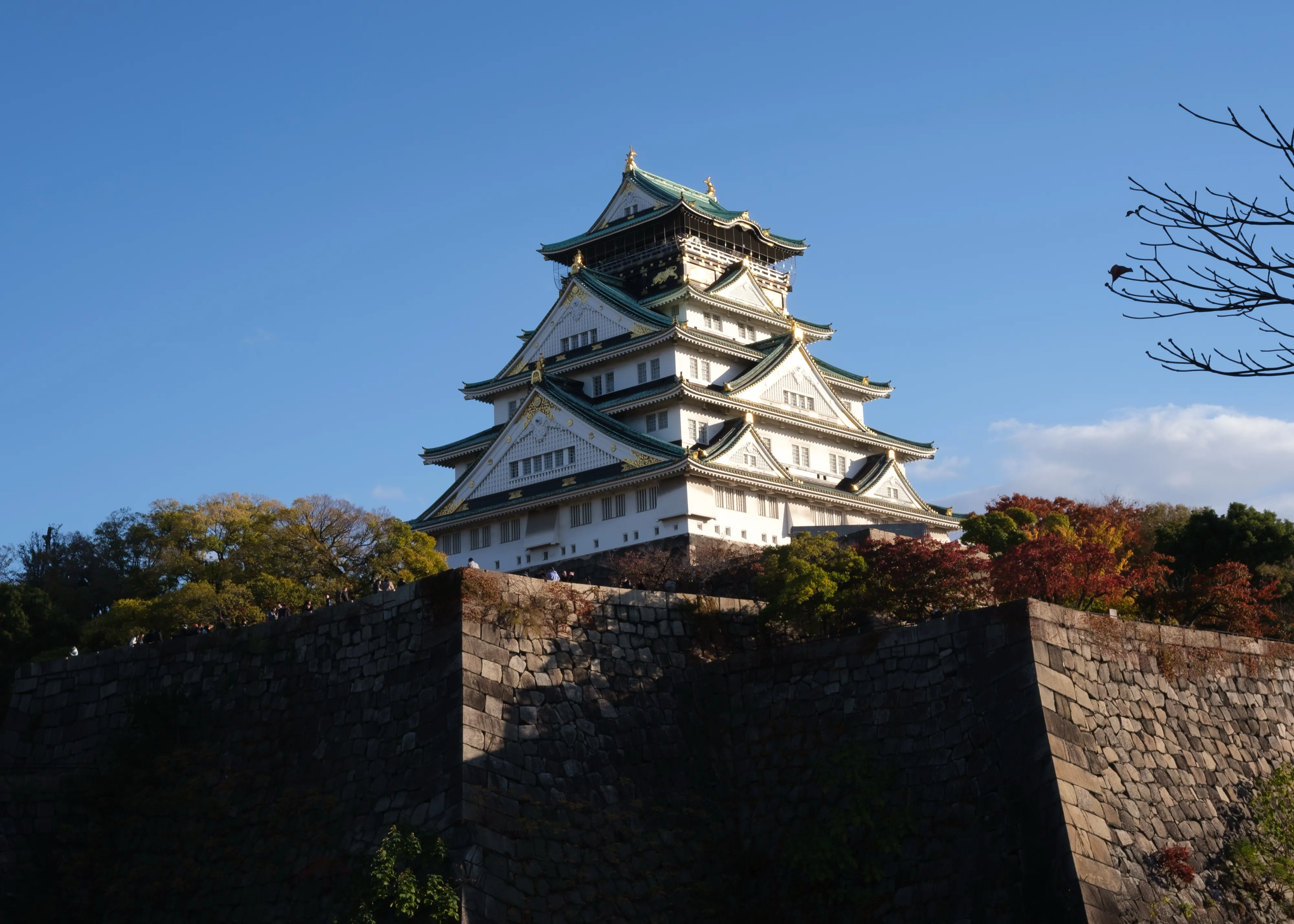 White multi-tiered Japanese castle with green roofs standing above a massive stone wall and autumn trees, against a deep blue sky