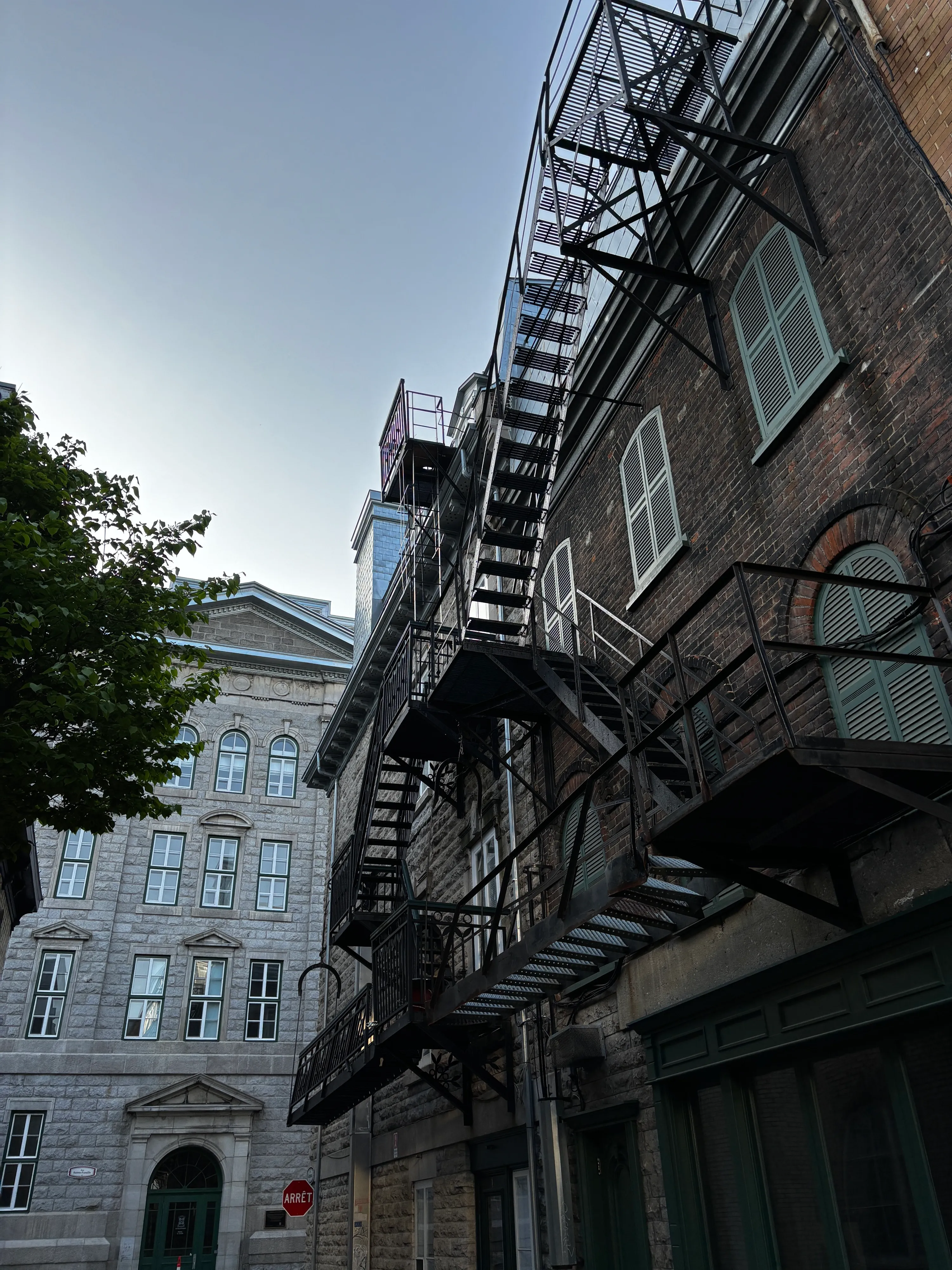 Zigzagging black fire escape climbing a brick building, with a grey stone building and a green tree beside it in Quebec City