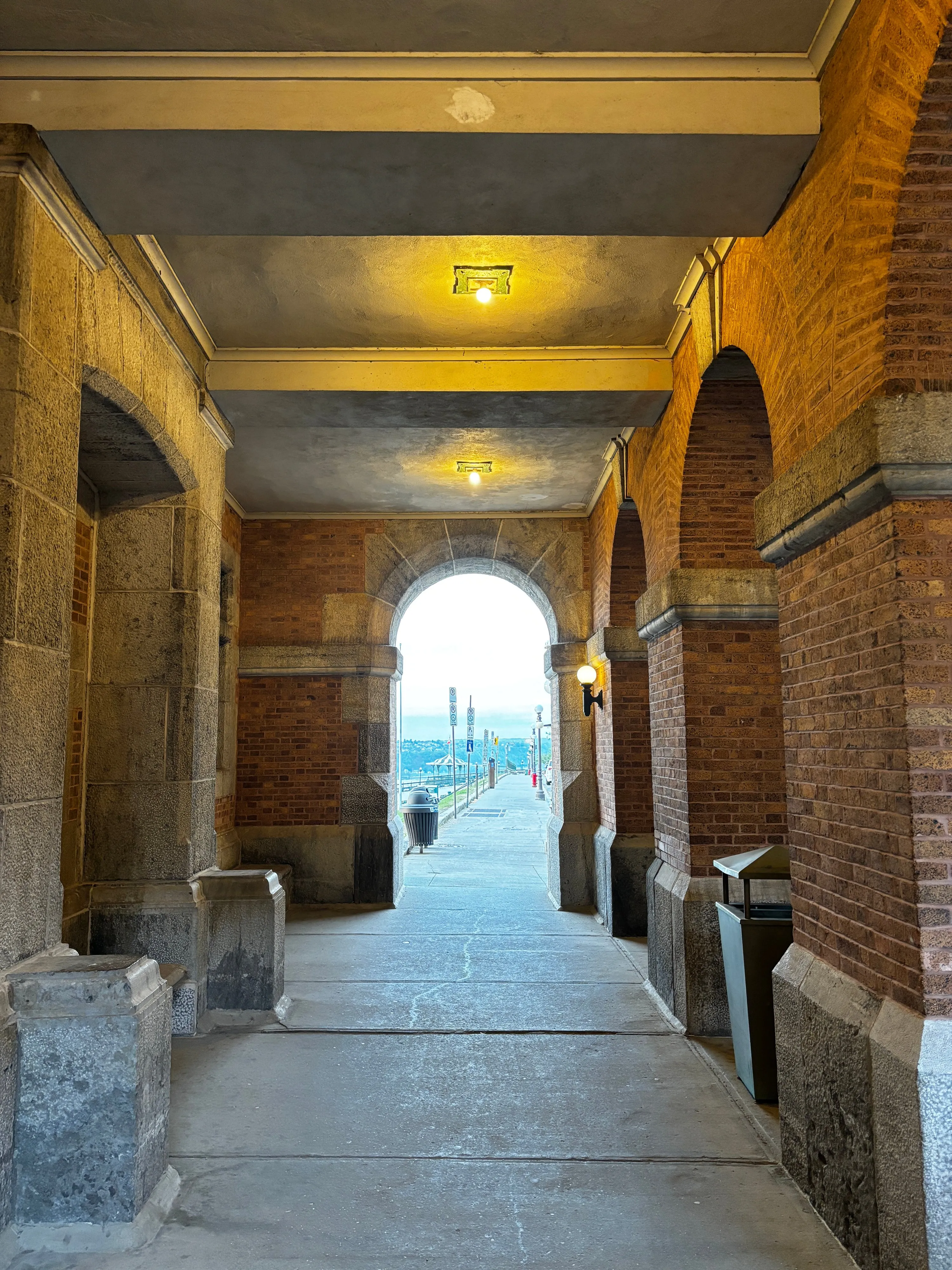 A brick and stone arcade framing a bright arched opening that looks out over a distant waterfront at dusk