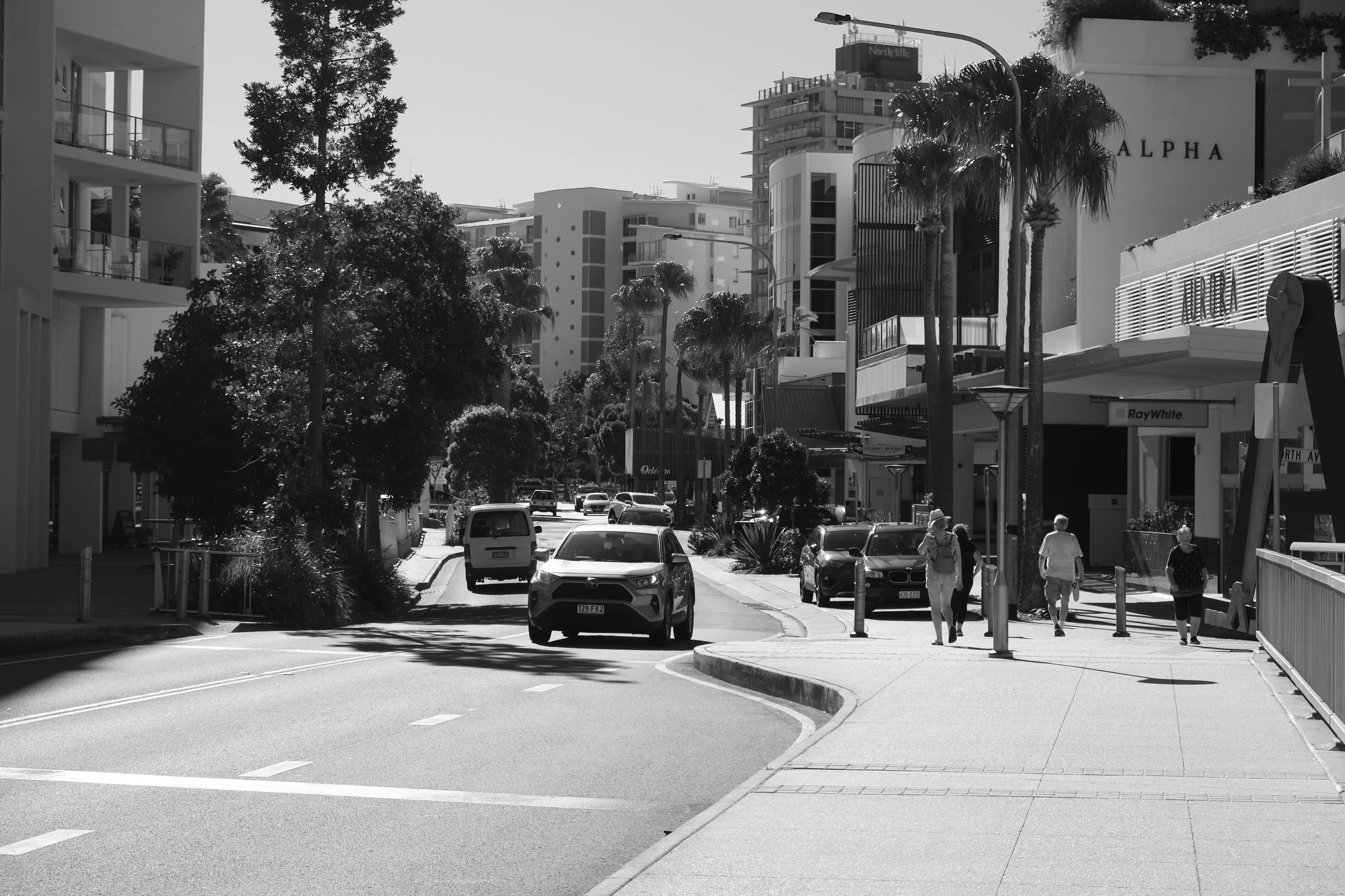 Seaside street with cars on the road and pedestrians along the footpath, lined with palms and apartment buildings in strong sunlight