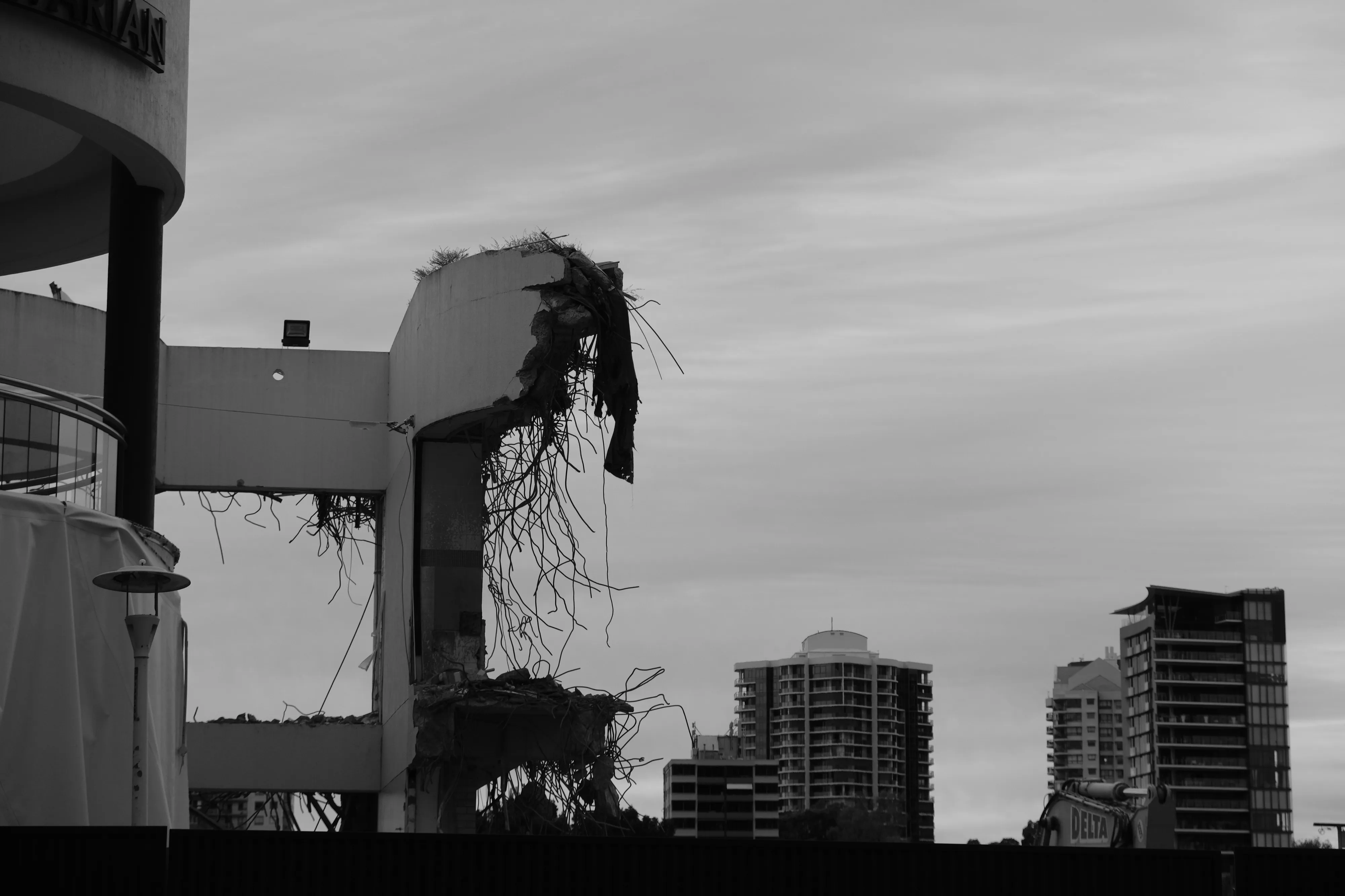 Partly demolished concrete building with exposed twisted steel reinforcing, set against a cloudy sky and apartment towers