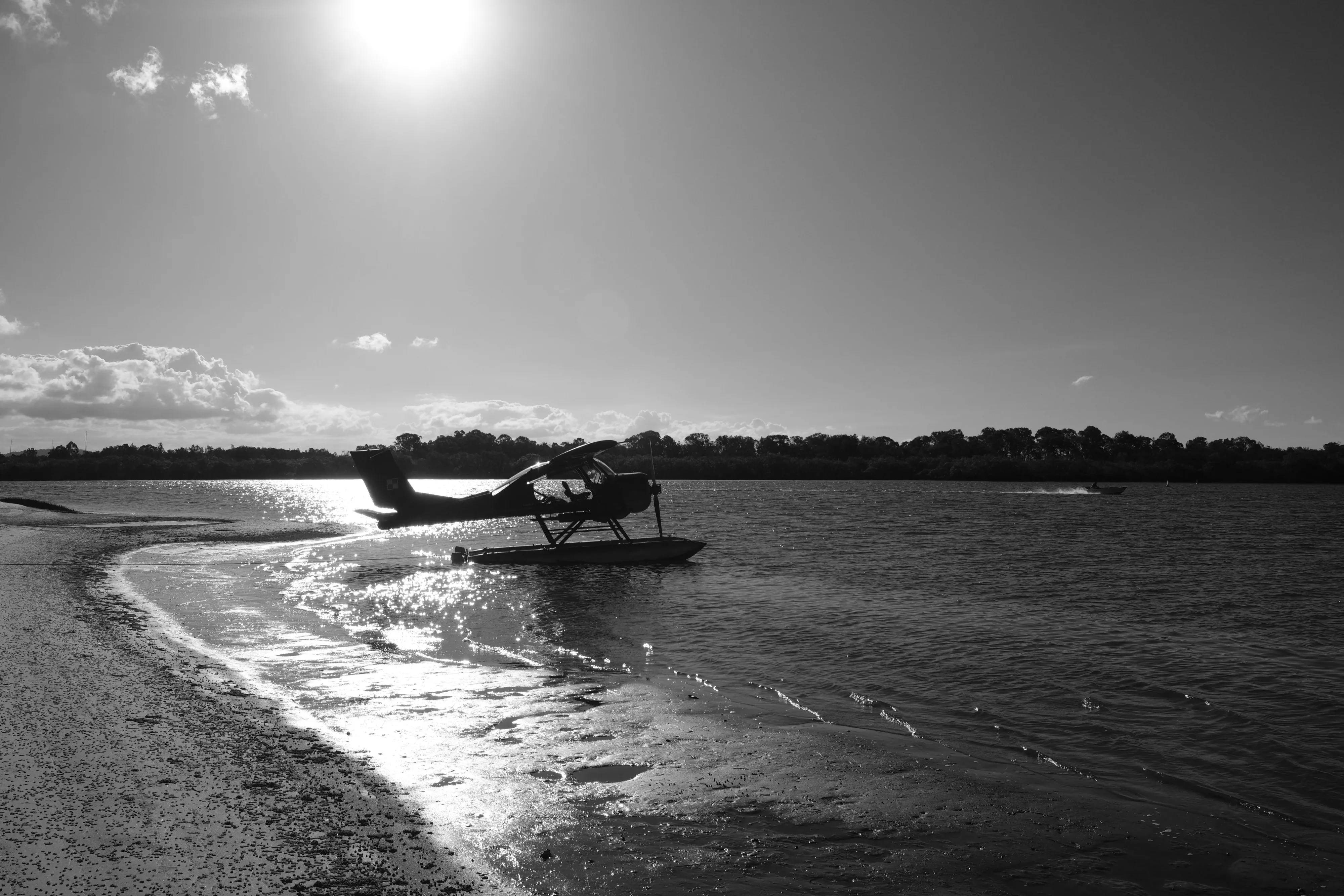 Small seaplane silhouetted at the water's edge against a bright sun, with ripples and wet sand in the foreground
