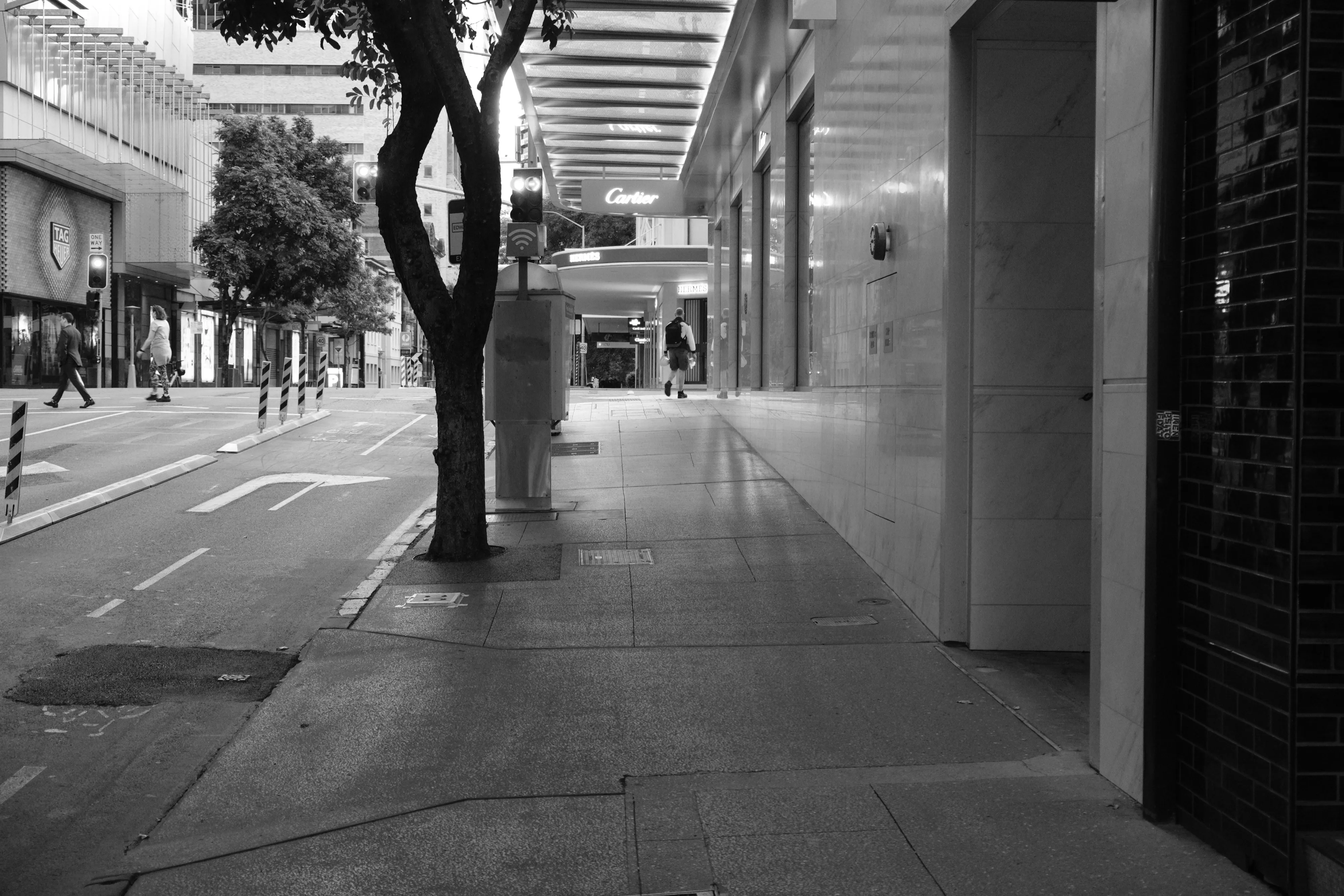 Empty city footpath beside a polished stone building, with a tree, a few distant pedestrians and a luxury shop sign in the background