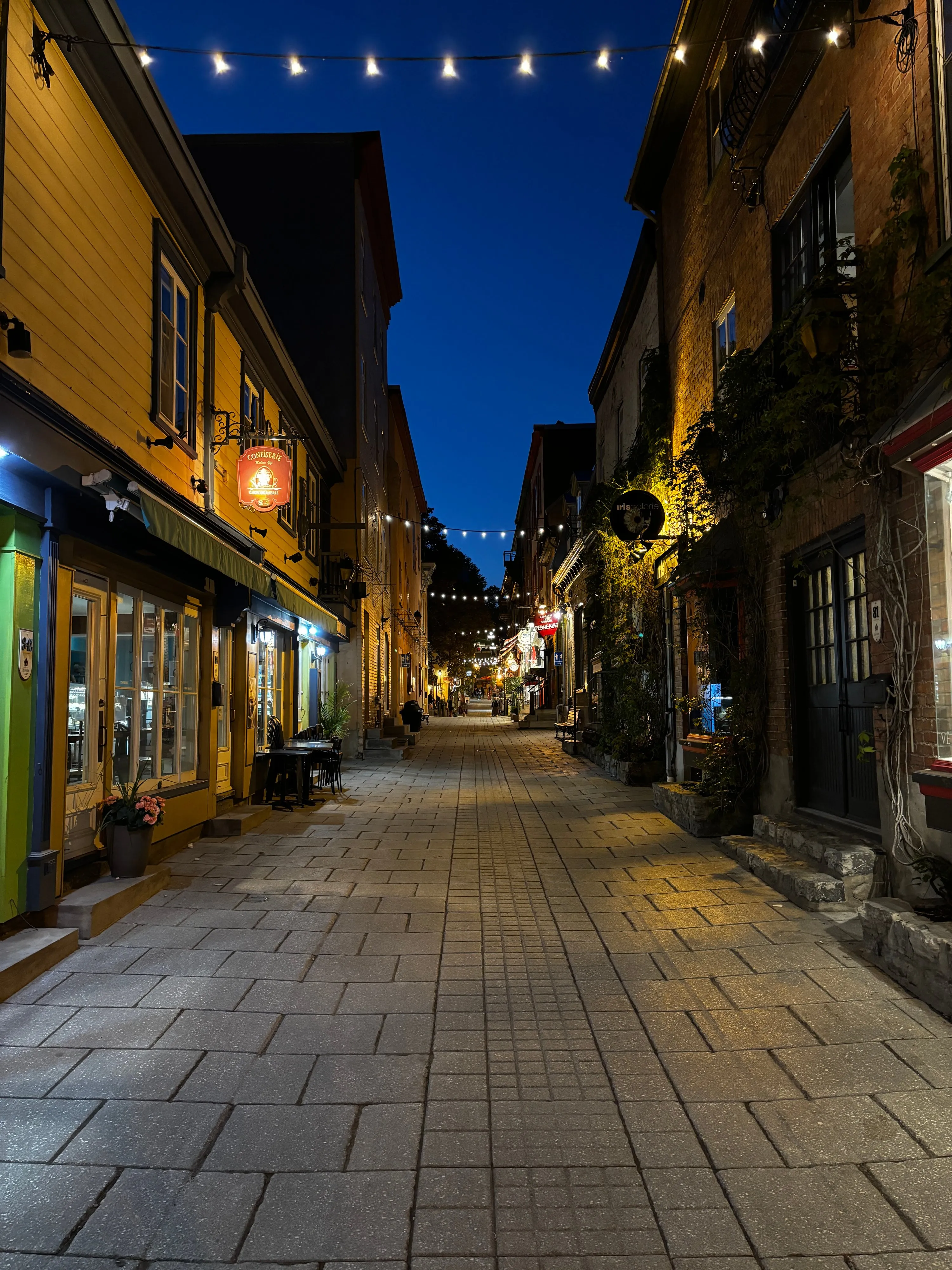 A cobbled street at night between yellow and brick buildings, with strings of lights overhead and a deep blue sky