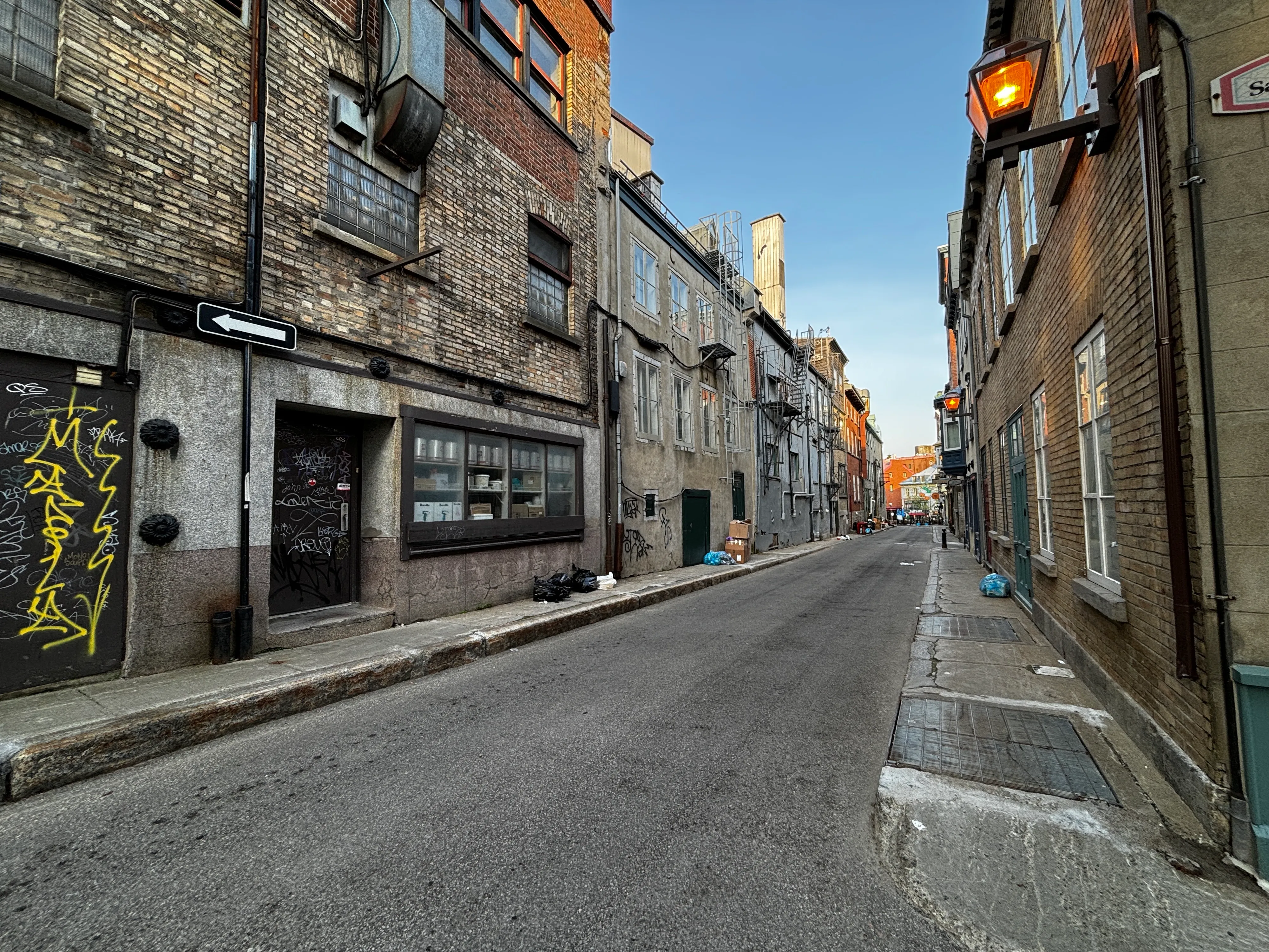 Narrow old stone street in Quebec City with a glowing lantern, graffiti on a doorway and tall brick buildings fading into the distance