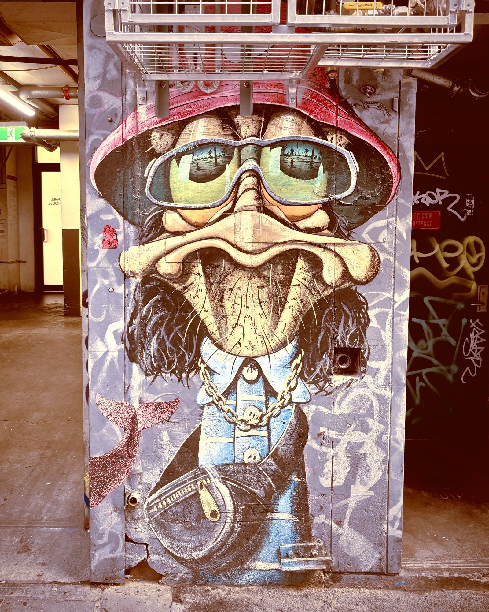 Large mural of a bearded character in a red cap and mirrored sunglasses painted across a laneway doorway, surrounded by tags
