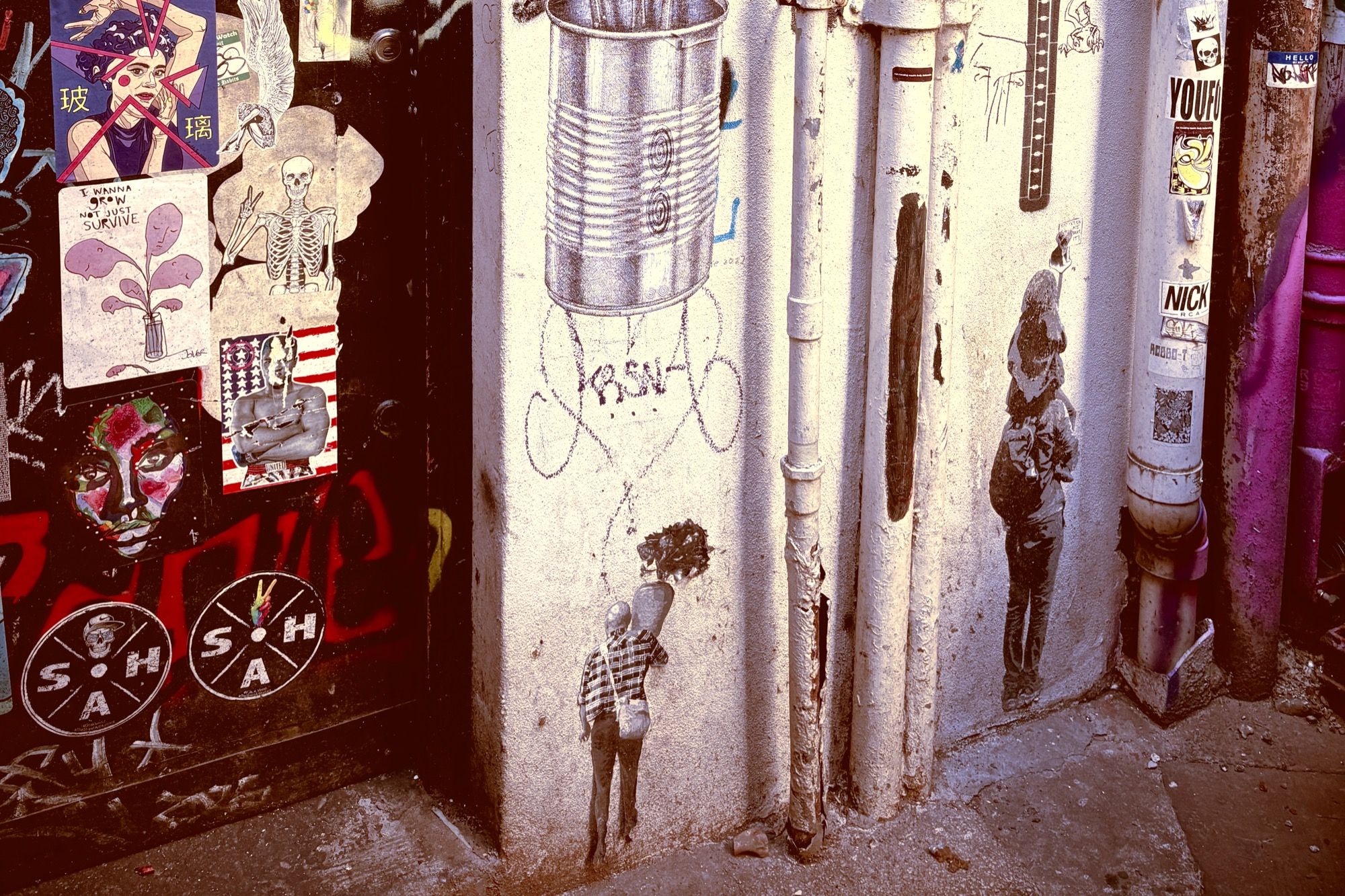 Laneway wall of stencil art and paste-ups, including a small stencilled figure holding a tin can drawn on the wall above